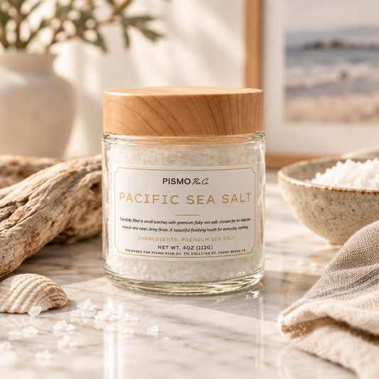 A clear glass jar of Pismo Sea Salt with a light wood-grain lid, set against a neutral gray background.